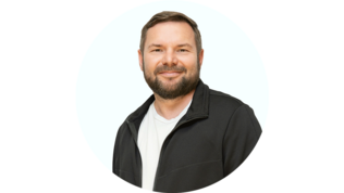 Ein Mann mit Bart lächelt freundlich in einem schwarzen Jackett vor einem hellen Hintergrund.