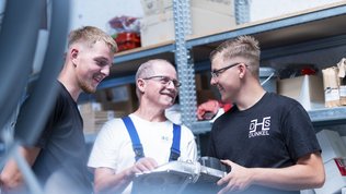 Drei Männer in einem Lagerraum lächeln und diskutieren über ein technisches Gerät, während sie sich gegenseitig anlächeln.