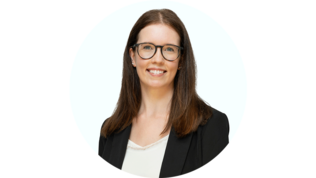 Eine lächelnde Frau mit langen, braunen Haaren trägt eine schwarze Blazerjacke und eine helle Bluse vor einem hellen Hintergrund.