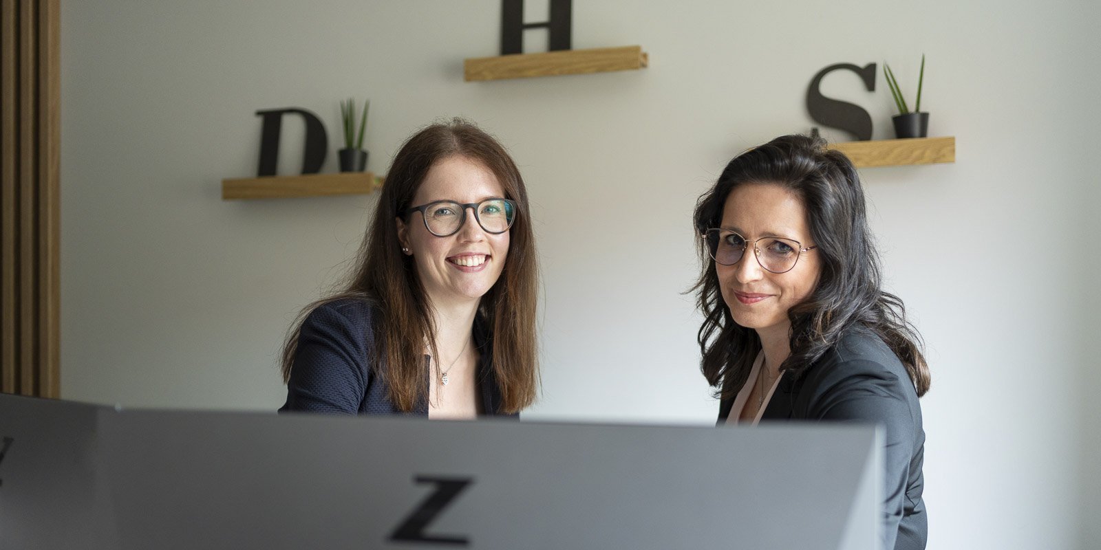 Zwei Frauen lächeln freundlich in einem modernen Büro, umgeben von Pflanzen und minimalistischer Dekoration.