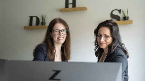 Zwei Frauen lächeln freundlich in einem modernen Büro, umgeben von Pflanzen und minimalistischer Dekoration.