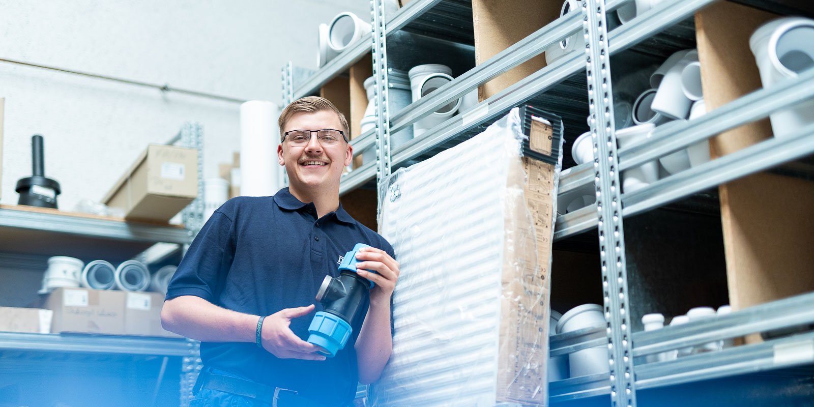 Ein junger Mann lächelt, während er ein blaues Rohrteil in einem Lager mit Regalen voller Rohre hält.