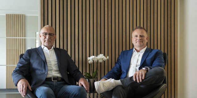 Zwei Männer sitzen entspannt in modernen Stühlen vor einer Holzvertäfelung, umgeben von einer eleganten Atmosphäre.