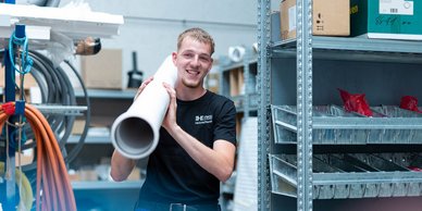 Ein junger Mann lächelt, während er ein großes Rohr in einem Lager mit Regalen voller Materialien hält.