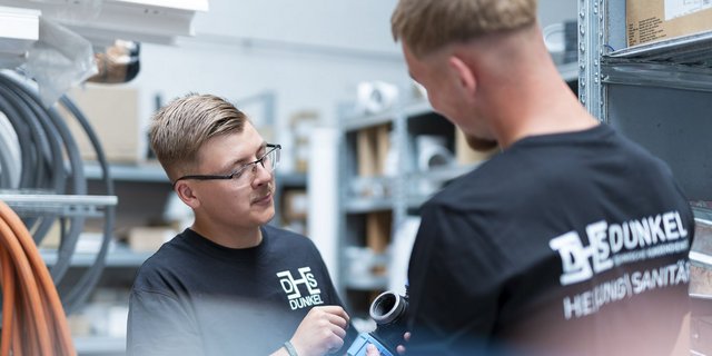 Zwei Männer in einem Lager diskutieren über Sanitärmaterialien, während sie auf eine blaue Rohrverbindung zeigen.