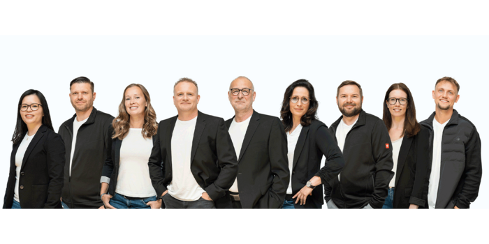 Eine Gruppe von neun Personen steht nebeneinander, alle tragen schwarze Blazer und weiße T-Shirts, vor einem hellen Hintergrund.