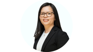 Eine lächelnde Frau mit langen, schwarzen Haaren trägt eine schwarze Blazerjacke und eine weiße Bluse vor einem hellen Hintergrund.