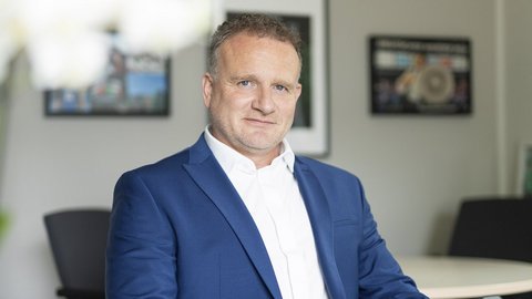 Ein Mann mit lockigem Haar trägt einen blauen Anzug und sitzt in einem modernen Büro mit Bildern im Hintergrund.