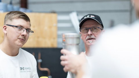 Ein junger Mann mit Brille hört aufmerksam einem älteren Mann mit Schnurrbart zu, der ein Metallrohr in der Hand hält.