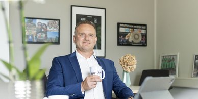 Ein Mann in einem blauen Anzug sitzt an einem Tisch und hält eine Tasse mit der Aufschrift 'BEARS' in der Hand.