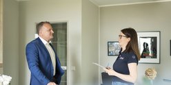 Ein Mann in einem blauen Anzug steht lächelnd in einem modernen Raum, während eine Frau mit Notizen ihm aufmerksam zuhört.