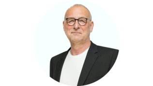 Ein Mann mit Brille und einem schwarzen Blazer lächelt leicht vor einem hellen Hintergrund.