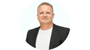 Ein Mann mit lockigem Haar trägt einen schwarzen Blazer über einem weißen T-Shirt und lächelt freundlich in die Kamera.