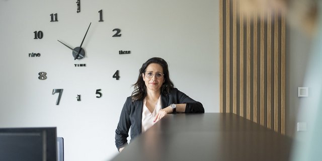 Eine Frau mit Brille steht an einem Tisch und lächelt, während eine große Uhr mit stilisierten Zahlen im Hintergrund sichtbar ist.