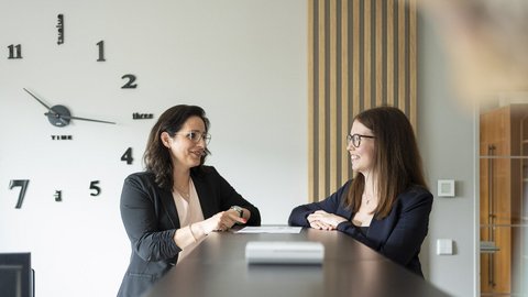 Zwei Frauen in formeller Kleidung unterhalten sich an einem modernen Empfangstresen, während eine große Wanduhr im Hintergrund sichtbar ist.
