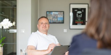 Ein Mann in einem weißen Hemd sitzt entspannt in einem modernen Büro und schaut freundlich in die Kamera.