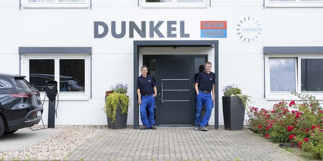 Zwei Männer in Arbeitskleidung stehen vor einem modernen Gebäude mit dem Schriftzug 'DUNKEL' und blühenden Pflanzen.