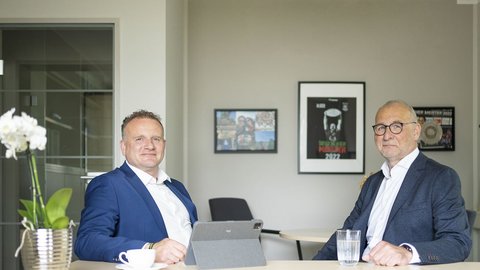 Zwei Männer in Anzügen sitzen an einem Tisch, umgeben von modernen Büromöbeln und dekorativen Bildern an der Wand.