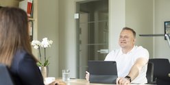 Ein Mann in einem weißen Polohemd lächelt, während er mit einer Person am Tisch spricht, umgeben von Pflanzen und Büchern.