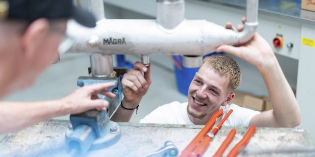 Ein junger Mann lächelt, während er an einem Metallrohr arbeitet, umgeben von Werkzeugen und einer hellen Werkstatt.