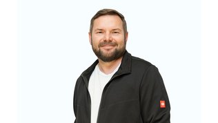 Der Mann mit einem freundlichen Gesichtsausdruck steht in einer schwarzen Jacke und strahlt Selbstbewusstsein aus.
