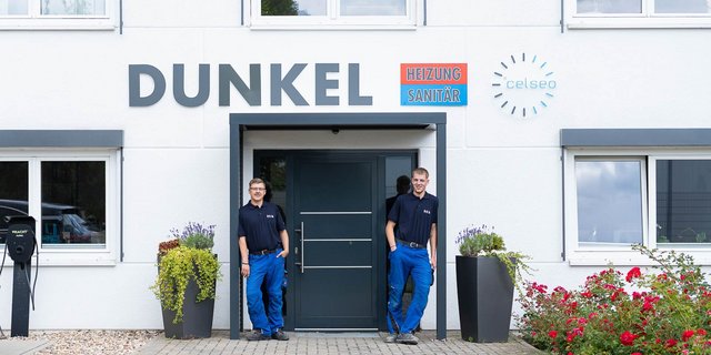 Zwei Männer in Arbeitskleidung stehen entspannt vor einem modernen Gebäude mit dem Schriftzug 'DUNKEL' und bunten Schildern.