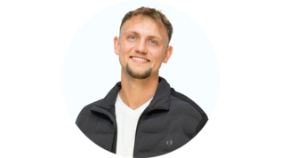 Ein junger Mann mit einem breiten Lächeln trägt eine schwarze Jacke und ein weißes T-Shirt vor einem hellen Hintergrund.