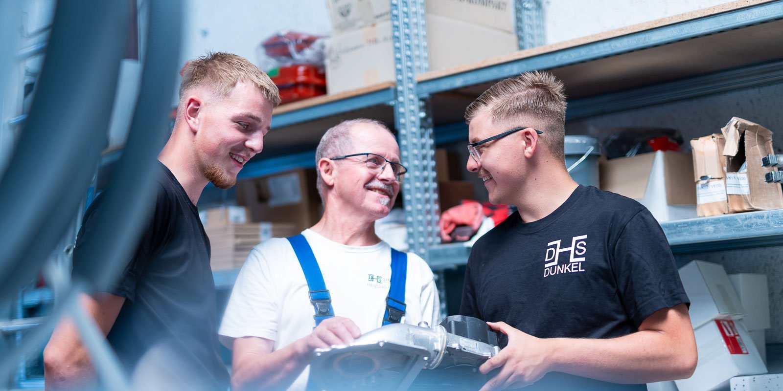 Drei Männer in einem Lager lächeln und diskutieren über ein Werkzeug, während Regale mit Materialien im Hintergrund sichtbar sind.