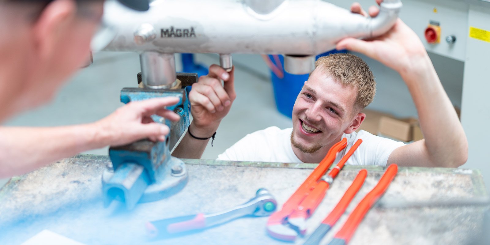 Ein junger Mann lächelt, während er an einem Metallrohr arbeitet, umgeben von Werkzeugen auf einer Werkbank.